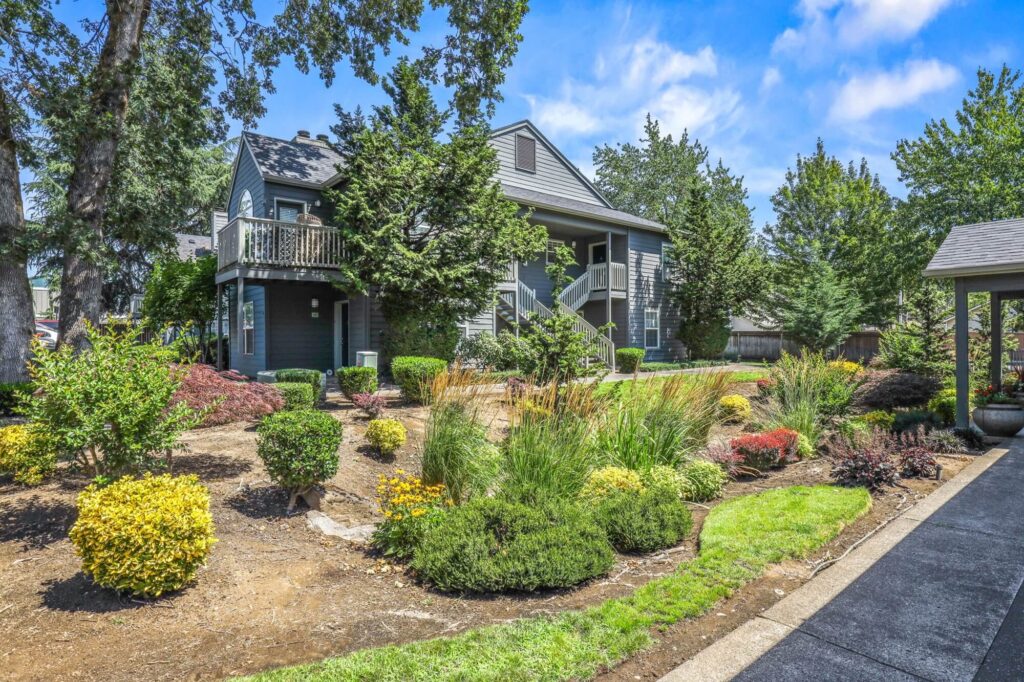 two story apartment building with exterior entrance and staircase to pathway, surrounded by lush foliage and landscaping
